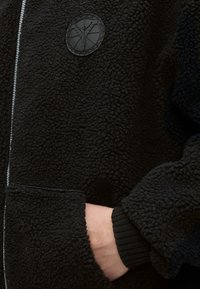 Χέρι σε τσέπη μαύρου fleece μπουφάν με υφή, φερμουάρ και στρογγυλό έμβλημα με φιγούρα με ανοιχτά χέρια.