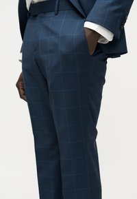 Άντρας που φοράει παντελόνια και σακάκι σε μπλε σκούρο καρό, με λευκό πουκάμισο, χέρια στις τσέπες, στέκεται μπροστά σε απλό φόντο.