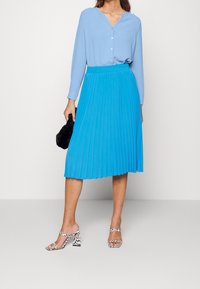 Gonna plissettata blu con vita alta, abbinata a una camicia leggera azzurra con bottoni. Accentuata da una pochette in pelliccia sintetica nera e tacchi con motivo.