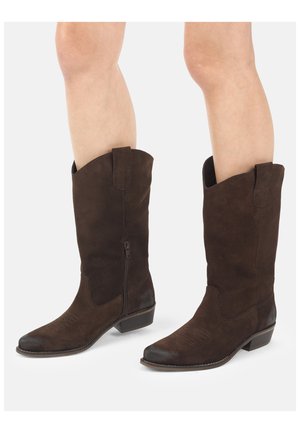 Ben iført mørkebrune ruskindskaubojstøvler til midt på læggen med lave hæle, lynlåse i siden og synlige sting på tåen på en lys baggrund.