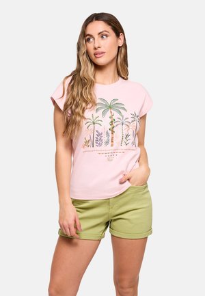 Joven mujer con cabello largo y ondulado, vistiendo una camiseta rosa claro con palmeras bordadas y pantalones cortos verdes remangados, con la mano en el bolsillo.