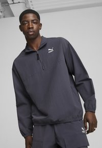 Puma lagana jakna sa patentnim zatvaračem u tamnosivoj boji; sadrži visoki ovratnik, duge rukave, elastične manžete i detalj s logotipom na grudima.