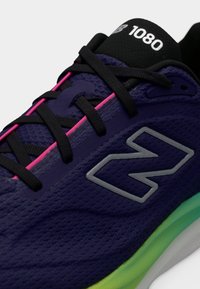 Μωβ αθλητικό παπούτσι New Balance 1080 με μαύρα κορδόνια, ροζ λεπτομέρειες στη θηλιά των κορδονιών, ανακλαστικό λογότυπο "N" και πράσινη σόλα με διαβάθμιση.