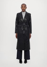 Μαύρο δερμάτινο παλτό τριάντα με ζώνη στη μέση, μακριά μανίκια, γιακάς notch και δύο μπροστινές τσέπες, φορεμένο πάνω από γκρι ζιβάγκο και παντελόνι με φαρδιά πόδια.