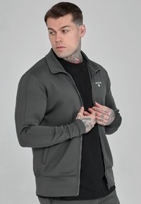 Zip-up jacket in dark gray, made of smooth fabric. Features a collar, side pockets, and a small logo on the chest. Fitted design.