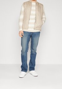 Man som bär en beige vadderad väst över en vit randig tröja, blå jeans och vita sneakers, stående mot en vit bakgrund.