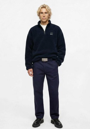 Ung mand med blondt krøllet hår bærer en sort fleece-ziptrøje, navy bukser, sorte sko og en hvid T-shirt under trøjen.