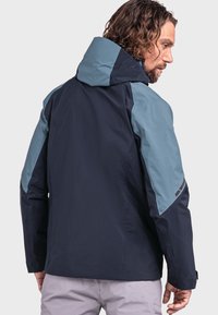 Wasserfeste Jacke mit navyblauem Körper, hellblauen Schultern, strukturiertem Stoff, verstellbarer Kapuze und reflektierenden Akzenten an den Ärmeln.