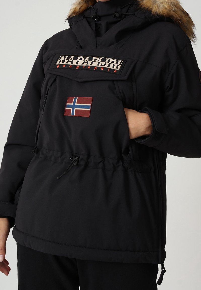 Veste d'hiver noire Napapijri avec capuche en fourrure, main dans la poche avant, et écusson du drapeau norvégien sur la poitrine.