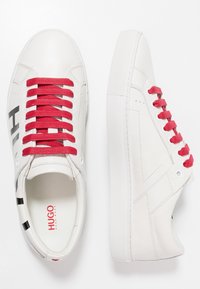 Baskets en cuir blanc avec des lacets rouges, une texture lisse et des détails de logo noirs sur le côté. Semelle en caoutchouc plate avec un motif. 