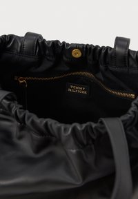 Εσωτερικό μαύρης δερμάτινης τσάντας tote με συγκεντρωμένη κορυφή, που διαθέτει τσέπη με χρυσό φερμουάρ και ετικέτα που αναγράφει "TOMMY HILFIGER." Λείος υφής.