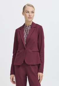 Žena u burgundy sakou i hlačama, nosi kariranu košulju i gleda desno, na jednostavnoj bijeloj pozadini.