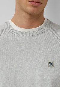 Jasnoszary sweter o dzianinowej strukturze z prążkowanym okrągłym dekoltem i małą naszywką z logo na piersi. Faktura wydaje się miękka i gładka.