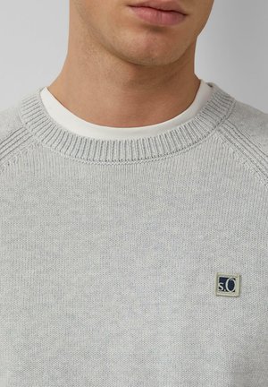 Maglione grigio chiaro lavorato a maglia con colletto rotondo a coste e una piccola toppa con logo sul petto. La texture sembra morbida e liscia.