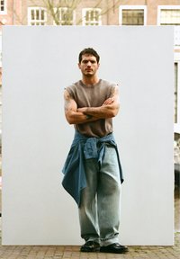 Muž s tetováním, který má na sobě nátělník a široké džíny, modrou kostkovanou košili svázanou kolem pasu, stojící se založenýma rukama.