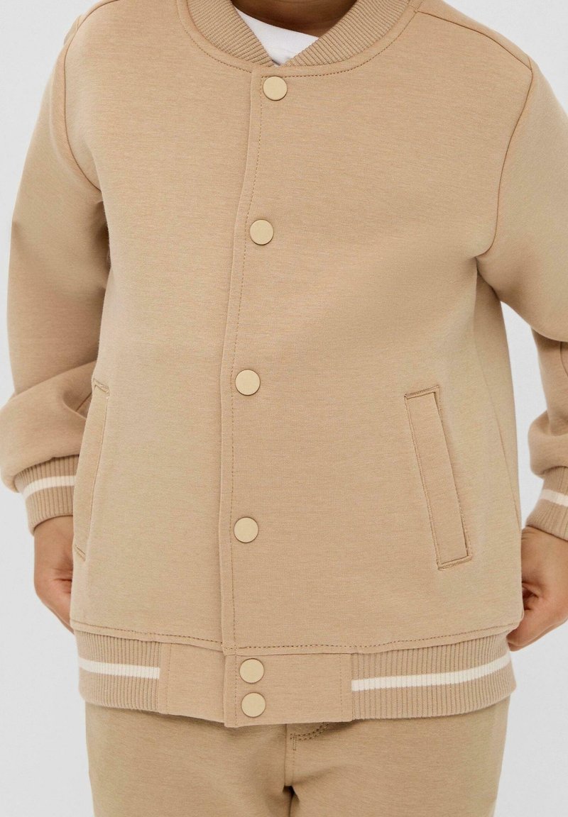 Enfant portant une veste beige boutonnée avec poignets et ourlet côtelés, avec deux poches avant et des détails rayés blancs sur les poignets et l'ourlet.