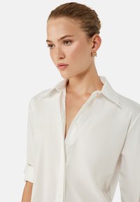 Joven mujer con cabello rubio recogido en un moño, vistiendo una camisa blanca de botones y pendientes de aro dorados, mirando pensativamente hacia un lado.