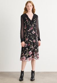 Schwarzes Blumenkleid mit Rüschenausschnitt, langen durchsichtigen Ärmeln und einem fließenden Rock, das mit rosa und weißen Blumen verziert ist, getragen mit schwarzen Knöchelstiefeln.