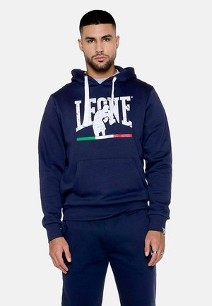 Homme portant un sweat à capuche Leone bleu marine avec des cordons blancs et un pantalon assorti, debout devant un fond clair uni.
