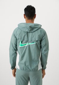 Kerge türkiissinine tuulejakker, millel on kapuuts, valge külgpaneel ja peegeldav roheline Nike logo seljal.