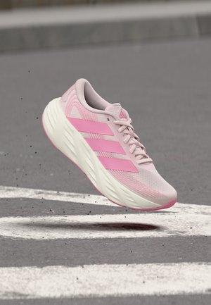 Zapatilla de correr rosa claro con tres rayas rosa más oscuras, suela blanca y cordones, flotando sobre una carretera de asfalto con marcas blancas.