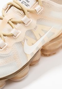 Baskets Nike avec une empeigne translucide beige, des motifs texturés, des lacets crème et un swoosh blanc, le tout reposant sur une semelle chunky semi-transparente.