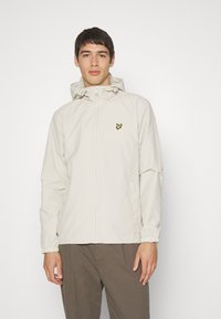Beige kevyt takki, jossa on vetoketjuluukku, säädettävä huppu, joustavat hihat ja pieni keltainen logo vasemmassa rinnassa.