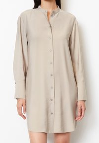 Beige Hemdblusenkleid mit langen Ärmeln, gefertigt aus glattem Stoff, mit hohem Kragen, durchgängigem Knopfverschluss und dezenten geknopften Manschetten.