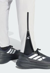 Białe sportowe spodnie na suwak z bocznymi otworami, w połączeniu z czarnymi butami z białymi paskami i teksturowaną podeszwą, noszone z czarnymi skarpetkami.