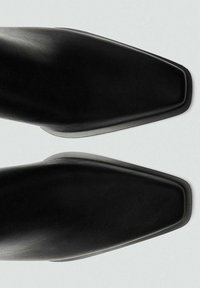 Chaussures en cuir noir avec un bout pointu et une finition lisse. Elles présentent un design minimaliste et une semelle robuste légèrement surélevée.