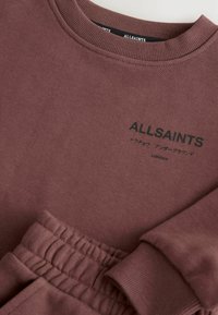 Mørkerød sweatshirt med ribbet rund hals, med sort logo tekst. Stoffet er glat og blødt, med matchende detaljer på ærmerne og taljen.