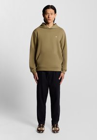 Olijfgroene hoodie met een buidelzak, gecombineerd met zwarte broek en gestreepte bruine schoenen, met een subtiele logo op de borst.