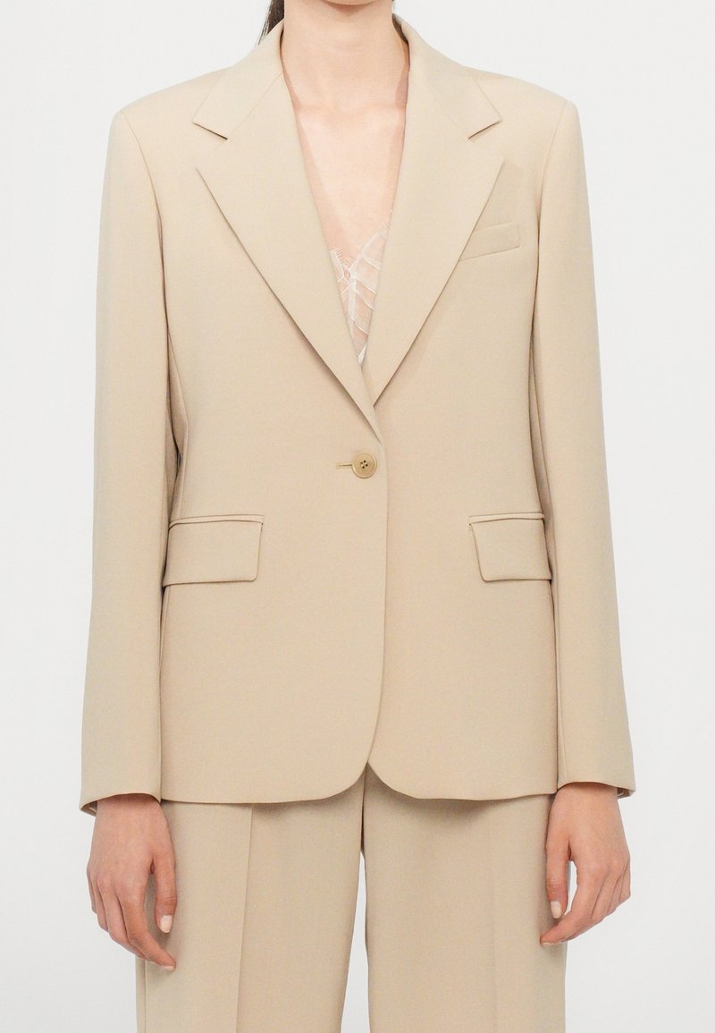 Blazer beige ajusté avec fermeture à un bouton porté sur un top en dentelle, assorti à un pantalon beige.
