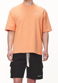 Hombre vistiendo una camiseta de manga corta suelta de color naranja y pantalones cortos cargo negros con cordones blancos, ambos de la marca "PEGADOR."