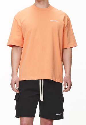 Hombre vistiendo una camiseta de manga corta suelta de color naranja y pantalones cortos cargo negros con cordones blancos, ambos de la marca "PEGADOR."