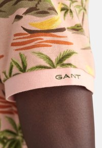 Krátký rukáv košile z růžové látky s tropickým vzorem ovoce a listů. Obsahuje zelené logo "GANT" na manžetě.