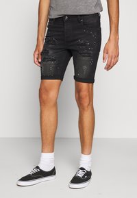 Shorts en denim noir avec déchirures et clous, ourlets retroussés et coupe ajustée. Portés avec des baskets noires et des chaussettes blanches.