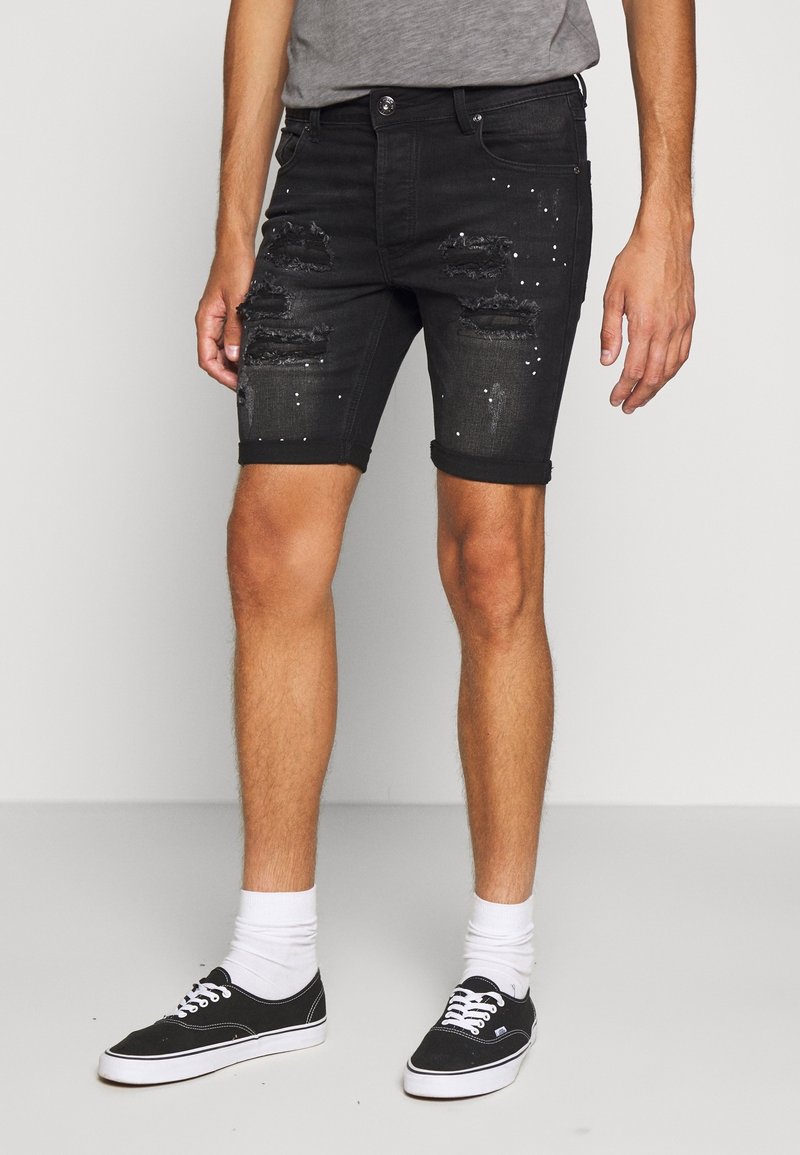 Shorts en denim noir avec déchirures et clous, ourlets retroussés et coupe ajustée. Portés avec des baskets noires et des chaussettes blanches.
