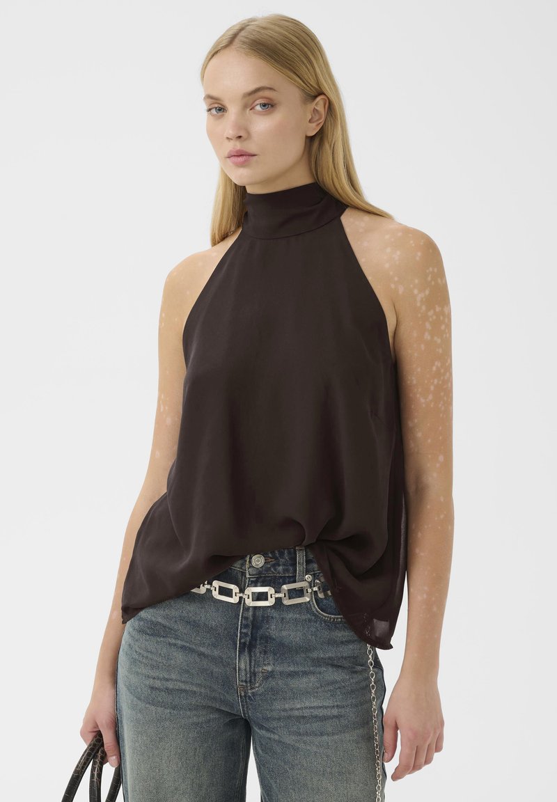 Femme blonde portant un haut noir sans manches à col halter, ceinture chaîne argentée, jean bleu, tenant un sac à main noir sur fond blanc uni.
