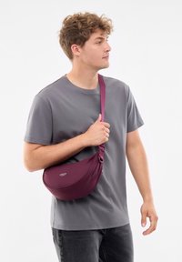 Borsa a tracolla bordeaux realizzata in materiale liscio, con una forma curva, tracolla regolabile e logo. Indossata a tracolla.