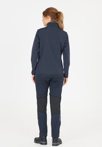 Marinblå softshell-jacka med hög krage, i kombination med marinblå och svarta byxor som har kontrasterande paneler och dragkedjor, samt svarta stövlar.
