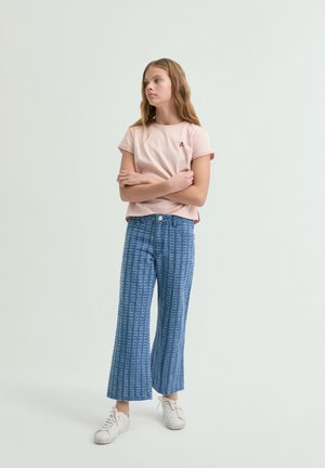 Une fille est debout, les bras croisés, portant un t-shirt rose clair, un jean large bleu à motifs, et des baskets blanches, sur un fond clair uni.