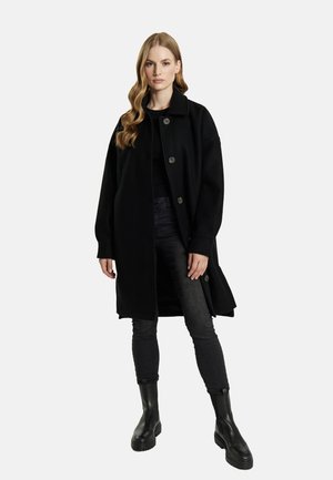 Frau mit langem, welligem Haar, die einen schwarzen, geknöpften Mantel, schwarze Hosen und schwarze Stiefeletten trägt, steht vor einem einfarbigen weißen Hintergrund.