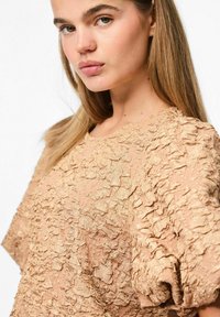 Top beige texturizzato con scollatura rotonda e maniche corte a sbuffo, che presenta una superficie increspata e sottili variazioni di tonalità.