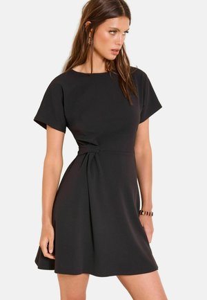 Frau trägt ein kurzärmeliges schwarzes Kleid mit seitlichem Knotendetail, steht mit einer entspannten Hand und schaut nach rechts.