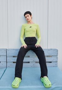 Világoszöld, hosszú ujjú crop top fekete Nike logóval, fekete jogger nadrággal és élénkzöld klumpával párosítva. Ül egy kék párnázott padon.