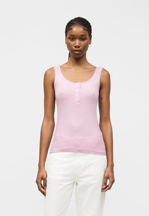 Γυναίκα με πλεξούδες, φοράει αμάνικο ανοιχτό ροζ μπλουζάκι με κουμπιά και λευκό παντελόνι, στέκεται μπροστά σε απλό ανοιχτό φόντο.