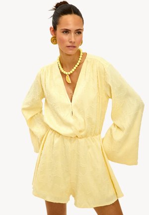 Rompers jaunes en tissu texturé avec un décolleté en V profond et des manches longues, doté d'une taille élastique et d'une coupe ample. Complété par des bijoux audacieux.