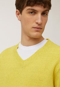 Geel V-halstrui met een gestructureerde brei-afwerking, voorzien van een klein logo op de borst, over een wit T-shirt.