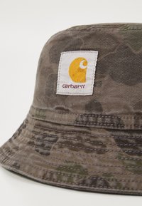 Bucket hat med camouflagemønster, lavet af tekstureret stof. Har et fremtrædende Carhartt-logo i gult og hvidt på siden.